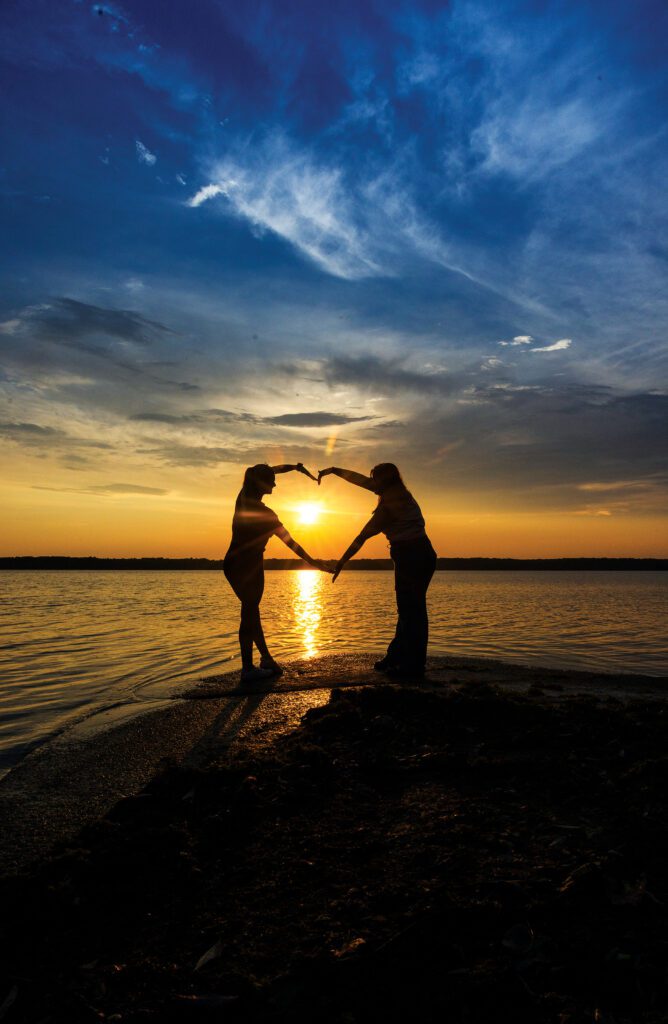 Two people forming a heart-shaped silhouette at sunset on the shoreline of Mosquito Lake State Park in Trumbull County, Ohio, with golden light reflecting off the water and a vibrant evening sky.