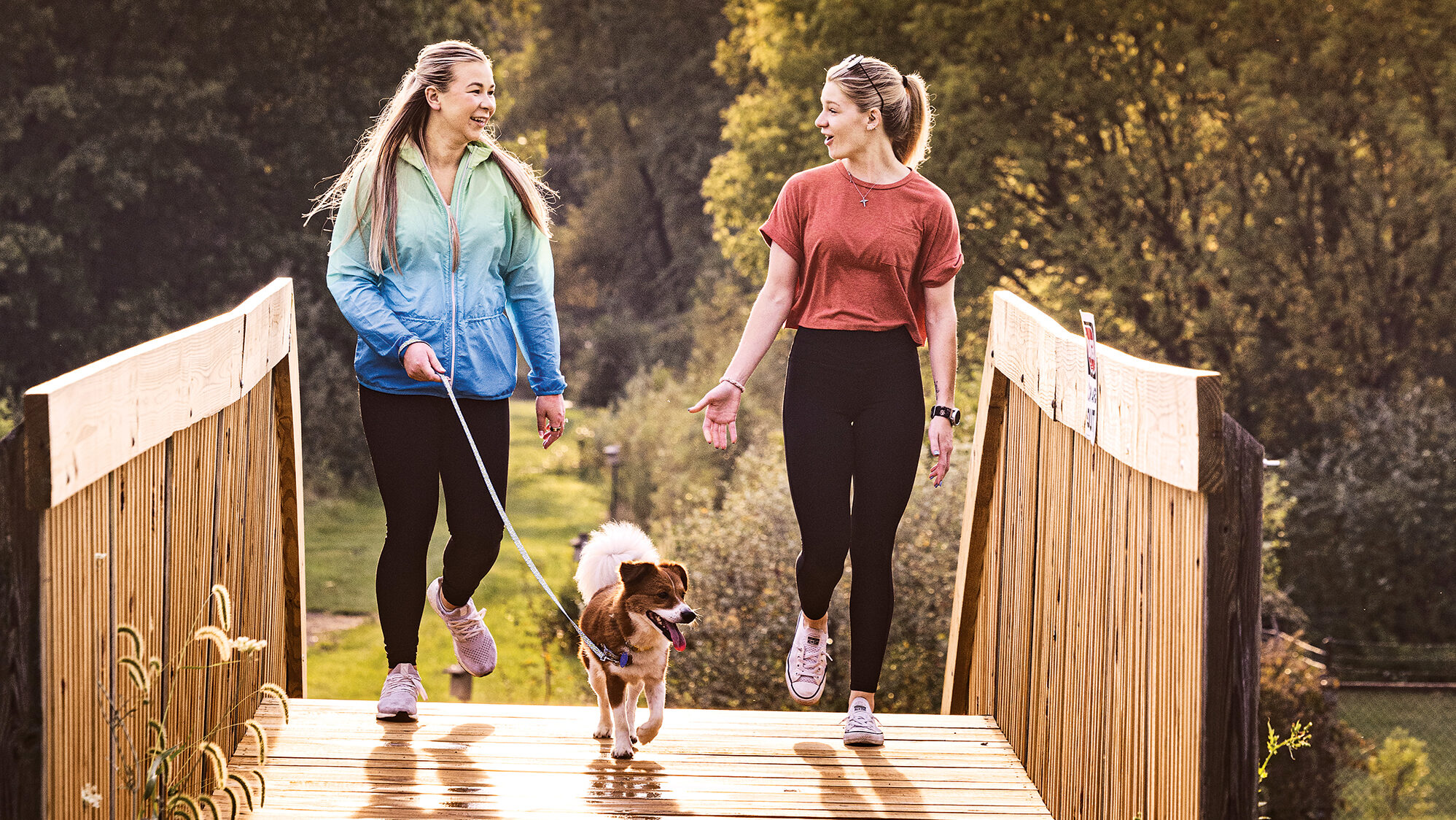 Two women walking a dog at sunset on Turkey Run Trail in Cortland, Ohio, surrounded by lush trees and nature in Trumbull County.