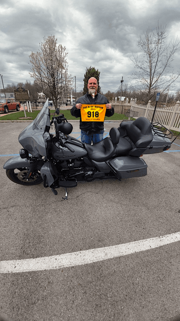 Motorcyclist holding 2025 Tour of Honor rider flag #918 in front of gray touring bike at Vietnam War Memorial in Warren, Ohio