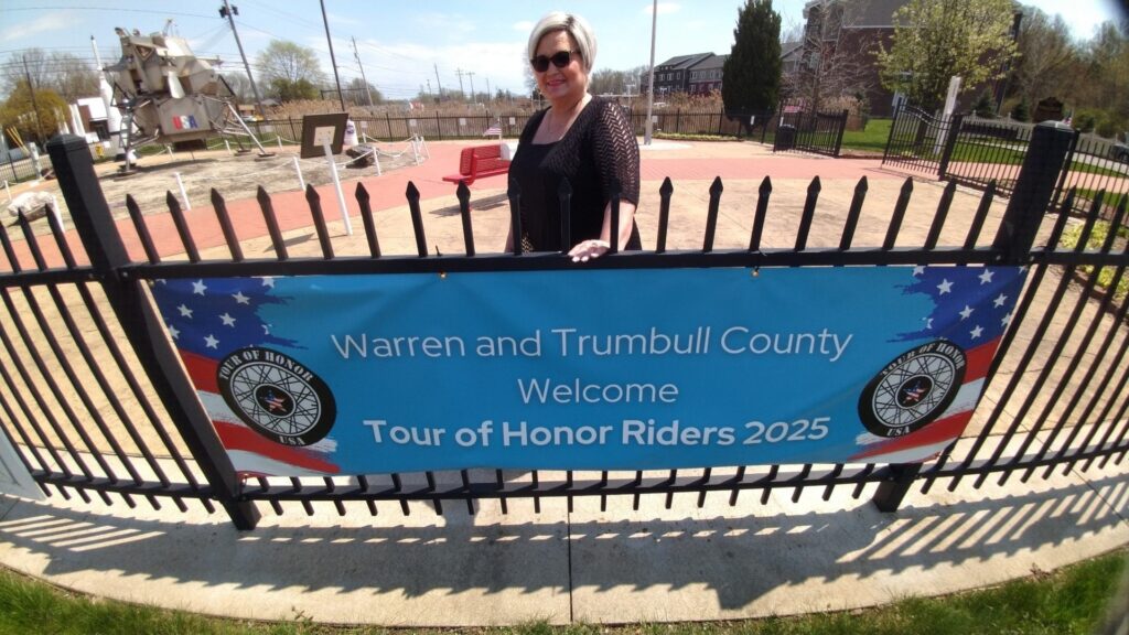 Linda Carpenter stands behind a banner welcoming 2025 Tour of Honor riders to Warren and Trumbull County, Ohio, near lunar lander display.
