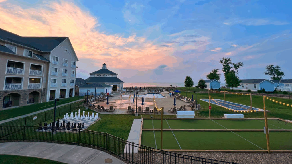 Sunset view of The Lodge at Geneva-on-the-Lake on Lake Erie, featuring the outdoor pool, string lights, bocce and shuffleboard courts, and nearby cottages in Ashtabula County, Ohio.